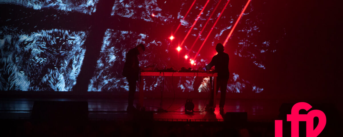 due ragazzi su un palco che suonano tramite tecnologie digitali con luci laser rosse dall'alto e videoproiezione sullo sfondo